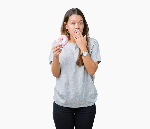 Young beautiful woman eating pink donut over isolated background cover mouth with hand shocked with shame for mistake, expression of fear, scared in silence, secret concept - Foto, immagini