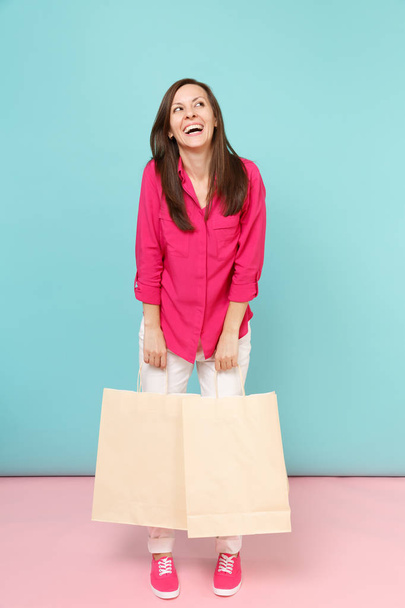 Full length portrait smiling young woman in rose shirt blouse, white pants hold shopping bags isolated on bright pink blue pastel wall background studio. Fashion lifestyle concept. Mock up copy space - Photo, Image