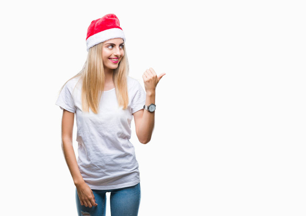 Young beautiful blonde woman christmas hat over isolated background smiling with happy face looking and pointing to the side with thumb up. - Photo, Image