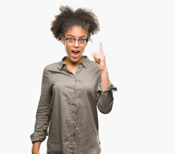 Joven mujer afroamericana con gafas sobre fondo aislado apuntando con el dedo hacia arriba con una idea exitosa. Salido y feliz. Número uno.
. - Foto, imagen