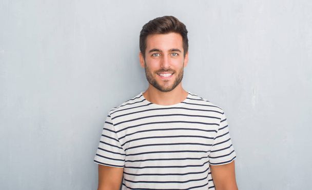 Csinos, fiatal férfi szürke grunge falon fárasztó sötétkék póló egy boldog és hűvös mosollyal arcán. Szerencsés személy. - Fotó, kép