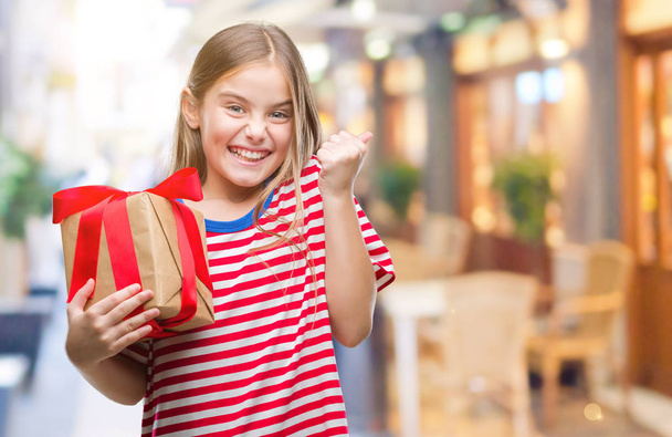 Young beautiful girl giving christmas or valentine gift over isolated background screaming proud and celebrating victory and success very excited, cheering emotion - Photo, Image