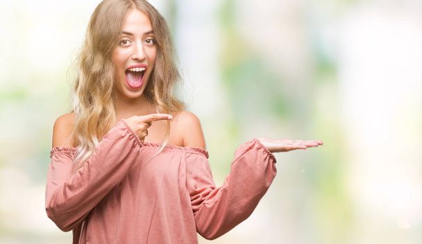 Beautiful young blonde woman over isolated background amazed and smiling to the camera while presenting with hand and pointing with finger. - Foto, afbeelding