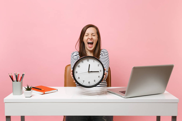 Jonge vrouw schreeuwen holding ronde wekker terwijl zitten, werken op witte Bureau met pc laptop geïsoleerd op pastel roze achtergrond. Carrière verwezenlijking bedrijfsconcept. Kopieer ruimte. Tijd loopt uit - Foto, afbeelding