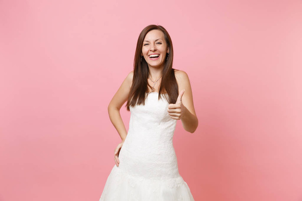Portrait of laughing joyful bride woman in elegant white wedding dress standing and showing thumb up isolated on pink pastel background. Wedding celebration concept. Copy space for advertisement - Photo, Image