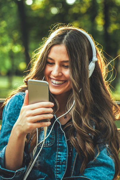 Όμορφη νεαρή χαμογελαστή κοπέλα κάθεται σε ένα παγκάκι πάρκου, ακούγοντας μουσική και κρατώντας ένα κινητό τηλέφωνο. Αναλαμπή φακού. - Φωτογραφία, εικόνα