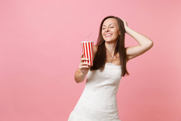 Portrait of smiling bride woman with closed eyes in white wedding dress keeping hand on head holding plastic cup with cola or soda isolated on pink pastel background. Wedding celebration. Copy space - Photo, Image