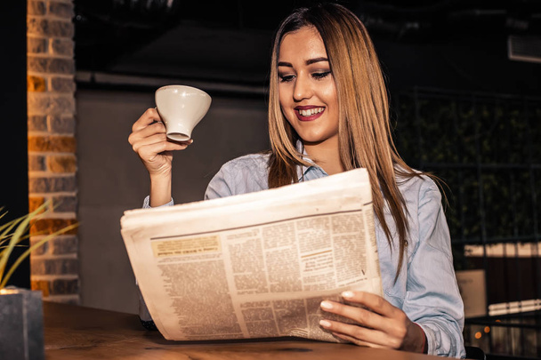 schöne junge Frau liest Zeitung und trinkt Kaffee, während sie morgens im Café Laptop benutzt.  - Foto, Bild