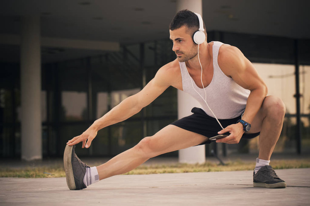 Young sportsman doing stretching exercise, preparing training in the park. While exercising, he listens to music via heasphones. Fitness, sport, lifestyle concept - Φωτογραφία, εικόνα