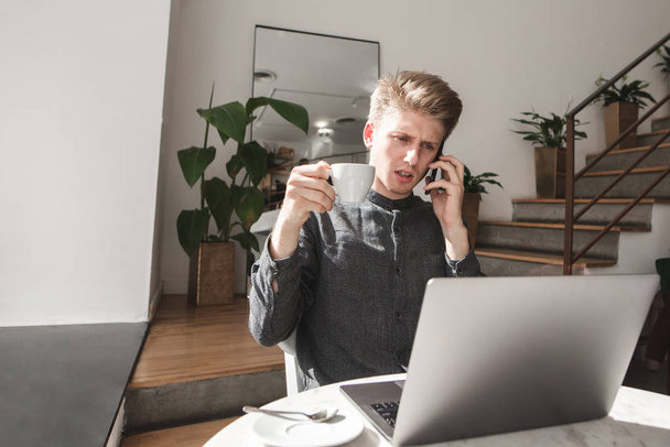 Ciddi genç adam rahat bir kafede bir akıllı telefon üzerinde konuşuyor, kahve içer ve dizüstü bilgisayar ekranında görünüyor. Konsantre genç işadamı bir kafede çalışıyor. - Fotoğraf, Görsel