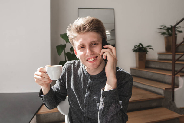 Estudiante habla por teléfono y bebe café.Primer plano retrato de un joven atractivo vestido con una camisa se sienta en un acogedor café, se comunica por teléfono, bebe café, mira la cámara y sonríe
 - Foto, Imagen