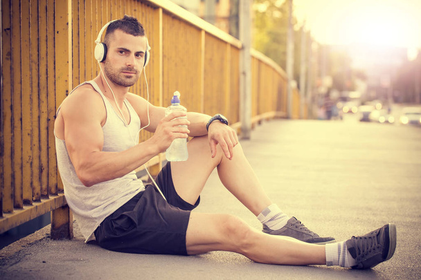 Portrait of young man drinking  water from a bottle while sitting, listening to music with headphones and resting after training. - Photo, Image