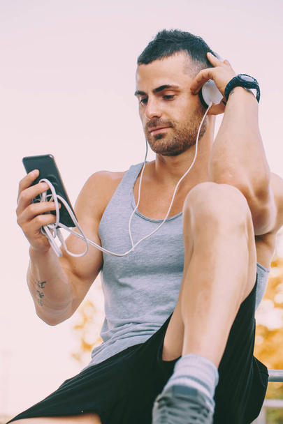 Resting after fitness. Young sportsman siting and listening to music with headphones after training outdoor. Fitness, sport, lifestyle concept - Foto, Bild