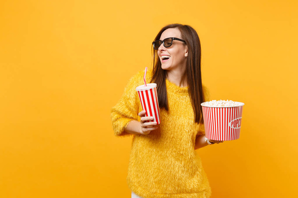 Радостная молодая женщина в 3D imax очки глядя в сторону смотреть фильм держать ведро попкорна, пластиковые чашки колы или газировки изолированы на желтом фоне. Люди искренние эмоции в кино, образ жизни
 - Фото, изображение