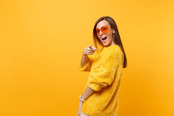Excited young woman in fur sweater orange heart glasses looking back pointing index finger on camera isolated on bright yellow background. People sincere emotions, lifestyle concept. Advertising area - Photo, image