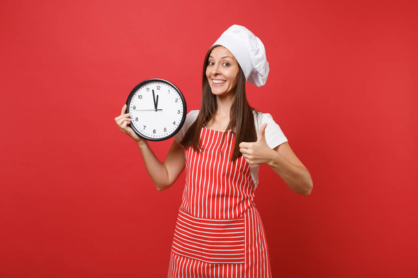 Huisvrouw vrouwelijke chef-kok of Bakker in gestreepte schort, wit t-shirt, toque koks hoed geïsoleerd op rode muur achtergrond. Lachende vrouw houdt in hand ronde klok haast omhoog. Bespotten kopie ruimte concept - Foto, afbeelding