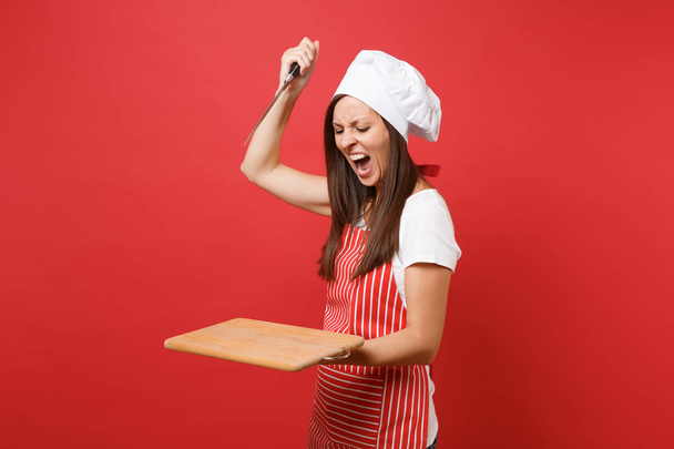 Femme au foyer cuisinière ou boulangère en tablier rayé blanc t-shirt, toque chefs chapeau isolé sur fond de mur rouge. Cris de colère femme tenir planche à découper en bois, couteau. Concept d'espace de copie simulé
 - Photo, image