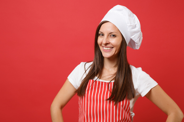 Huisvrouw vrouwelijke chef-kok of Bakker in rode gestreepte schort, wit t-shirt, toque koks hoed geïsoleerd op rode muur achtergrond. Portret van huishoudster brunette vrouw close-up. Bespotten kopie ruimte concept - Foto, afbeelding