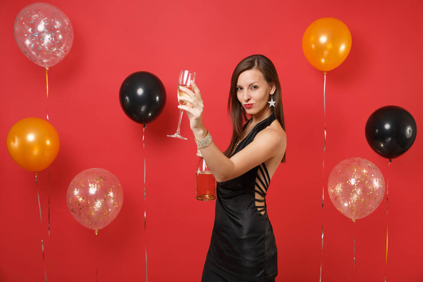 Stunning young girl in little black dress celebrating, holding bottle, rising hand with glass of champagne on bright red background air balloons. Happy New Year, birthday mockup holiday party concept - Photo, Image