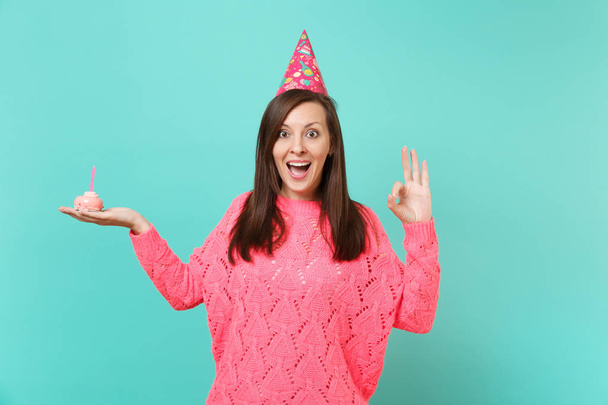 Excited woman in knitted pink sweater, birthday hat keeping mouth wide open, showing OK gesture hold in hand cake with candle isolated on blue background. People lifestyle concept. Mock up copy space - Photo, Image