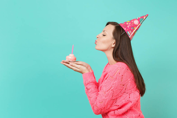 Vista lateral de la mujer joven en suéter de punto rosa, sombrero de cumpleaños soplando vela en la torta en las manos aisladas en azul turquesa retrato de estudio de fondo. Concepto de estilo de vida. Simular espacio de copia
 - Foto, imagen