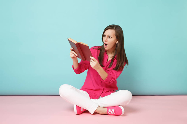 Ganzkörperporträt einer schockierten Frau in rosa Hemdbluse, weiße Hosen sitzen auf dem Boden lesen Buch isoliert auf rosa blau pastellfarbenen Wandhintergrund Studio. Mode-Lifestyle-Konzept. Attrappe Kopierraum - Foto, Bild
