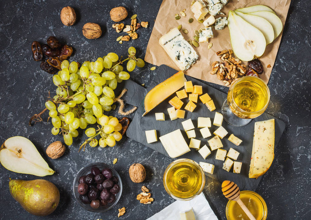 Различные сорта сыра на борту, оливки, фрукты, миндаль и бокалы вина на черном каменном столе. Вид сверху
 - Фото, изображение