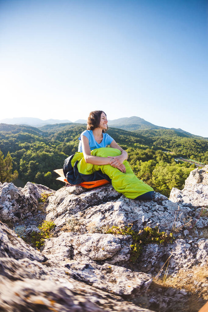 Kobieta siedzi w śpiwór o zachodzie słońca. Camping na szczycie góry. Uśmiechnięte dziewczyny odpoczywa po wspinaczki. Siedzi brunetka na siebie pompowania maty na tle gór. - Zdjęcie, obraz