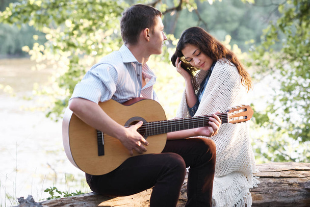 young couple sitting on a log by the river and playing guitar, summer nature, bright sunlight, shadows and green leaves, romantic feelings - Fotó, kép