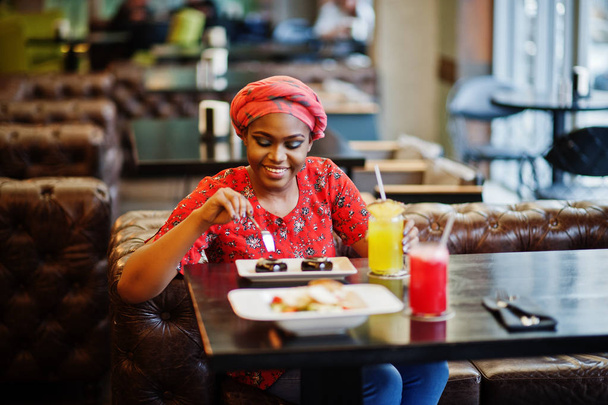 Stylish african woman in red shirt and hat posed indoor cafe, eat chocolate dessert cakes. - Fotografie, Obrázek