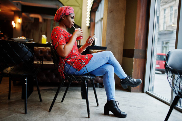 Stylish african woman in red shirt and hat posed indoor cafe and drinking strawberry lemonade with phone at hand. - Φωτογραφία, εικόνα