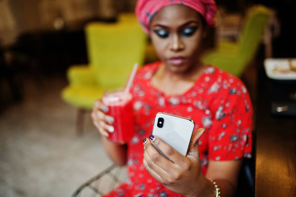 Elegante donna africana in camicia rossa e cappello posato caffè interno, bere limonata di fragole e fare selfie al telefono
. - Foto, immagini
