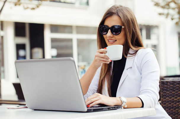 Счастливая молодая деловая женщина пьет кофе и пользуется ноутбуком в уличном кафе
 - Фото, изображение