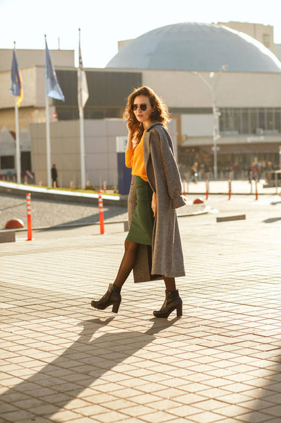 Atractiva mujer joven con el pelo rizado en gafas de sol con traje de moda y caminando por la calle soleada
 - Foto, Imagen