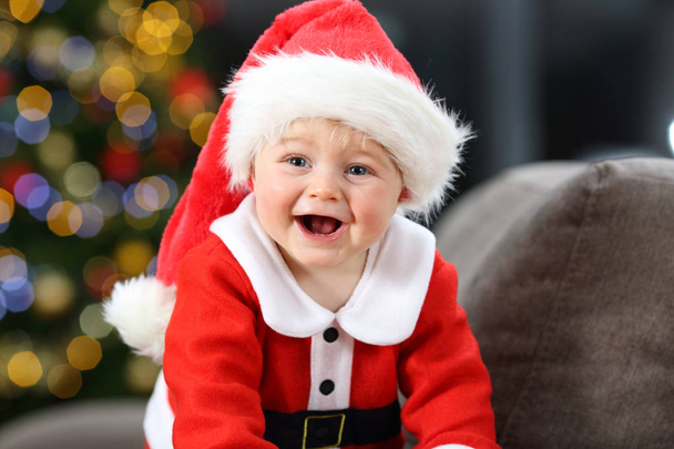 Joyful baby wearing a red santa clays disguise on a couch at home in christmas with a tree in the background - Photo, Image