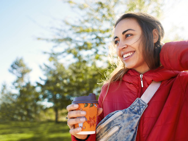 young woman in red jacket with blue jeans Waist bag drinking coffee outdoors - 写真・画像