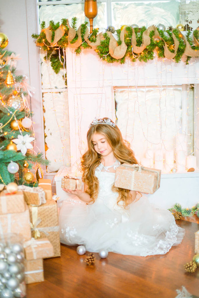 Cute little girl with long curly  hair at home near a Christmas tree with gifts and garlands and a decorated fireplace  - Photo, image