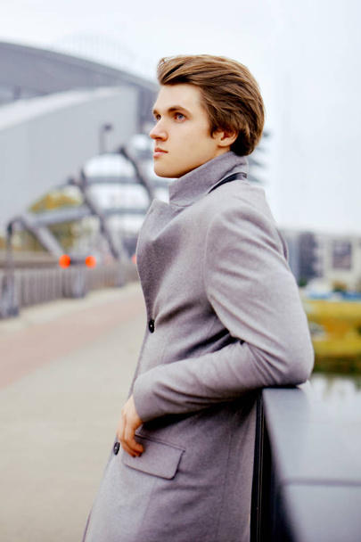 Young handsome man in coat. Fashionable well dressed boy smile in stylish coat. Boy outdoor at autumn. Elegant man with perfect hairstyle. Posing on the bridge. Fashion - Photo, Image