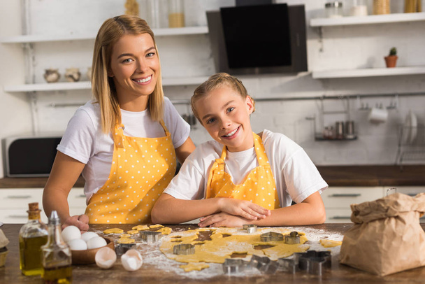 ευτυχισμένη μητέρα και κόρη στις ποδιές προετοιμασία τα cookies και να χαμογελά στη φωτογραφική μηχανή στην κουζίνα - Φωτογραφία, εικόνα