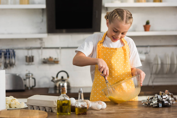 чарівний усміхнений хлопець у фартуху збиває яйця під час приготування їжі на кухні
 - Фото, зображення