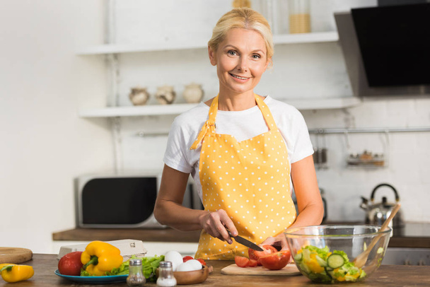 mutlu olgun kadın mutfakta yemek yaparken kameraya gülümseyen önlük - Fotoğraf, Görsel