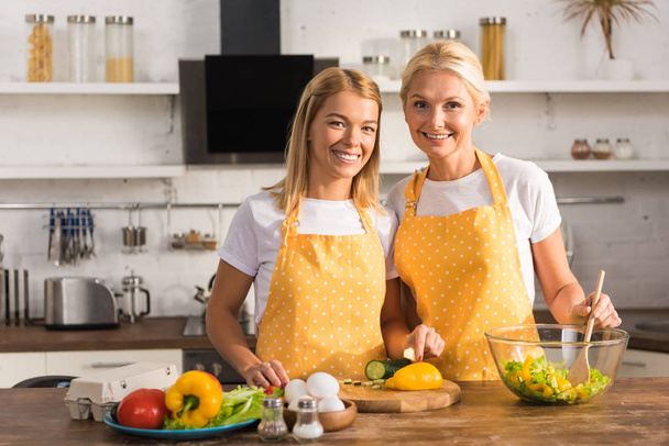 feliz madre adulta e hija en delantales sonriendo a la cámara mientras cocinan juntos
 - Foto, Imagen