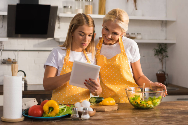взрослые мать и дочь в фартуках с помощью цифрового планшета во время приготовления пищи вместе
  - Фото, изображение
