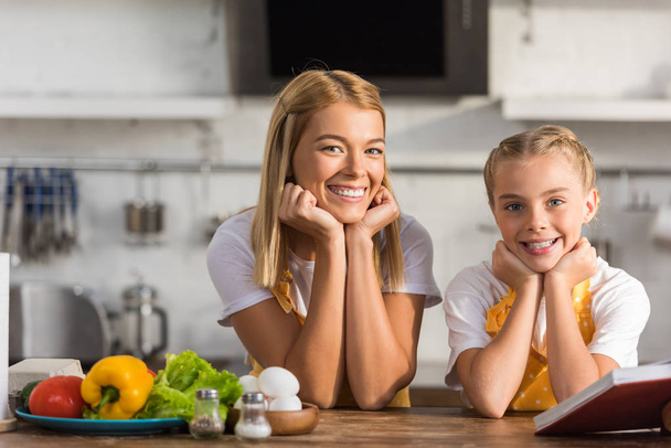 Glückliche Mutter und Tochter in Schürzen lehnen am Küchentisch und lächeln in die Kamera - Foto, Bild