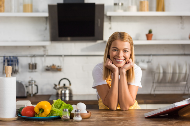 beautiful happy young woman in apron smiling at camera while cooking in kitchen  - Foto, afbeelding