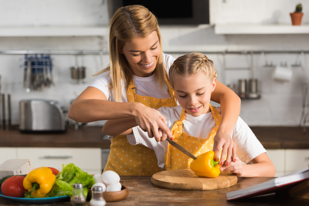 smiling young woman helping little daughter cutting pepper in kitchen - 写真・画像