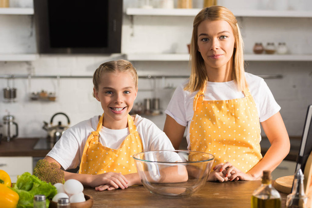 happy mother and daughter in aprons smiling at camera while cooking together  - Foto, immagini