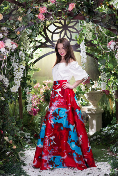 Mladé stylové ženy v luxusní červené hedvábí šaty s květinovým vzorem pózování pod kvetoucí obloukem - Fotografie, Obrázek