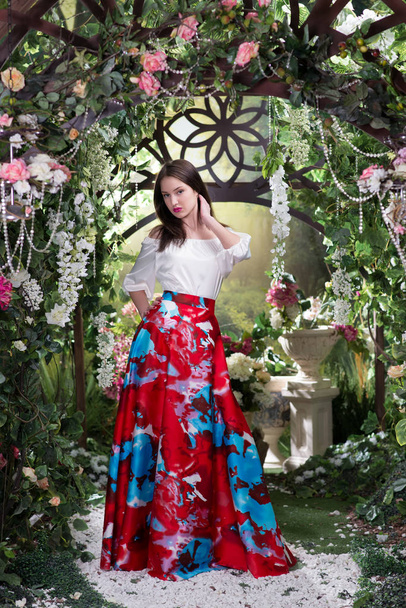 Молодая стильная женщина в роскошном красном шелковом платье с цветочным узором позирует под цветущей аркой
 - Фото, изображение