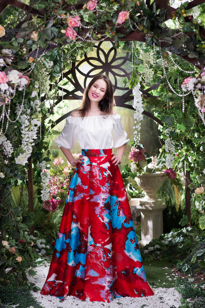 Mladé stylové žena v luxusní červené sukni s květinovým vzorem a hedvábnou blůzu pózování pod kvetoucí obloukem - Fotografie, Obrázek